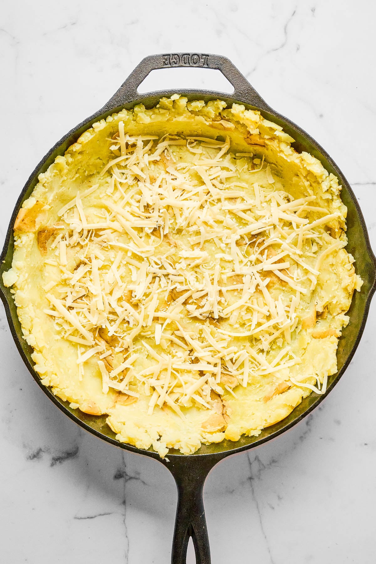 A cast iron skillet filled with a mashed potato mixture topped with shredded cheese, placed on a white marble surface.