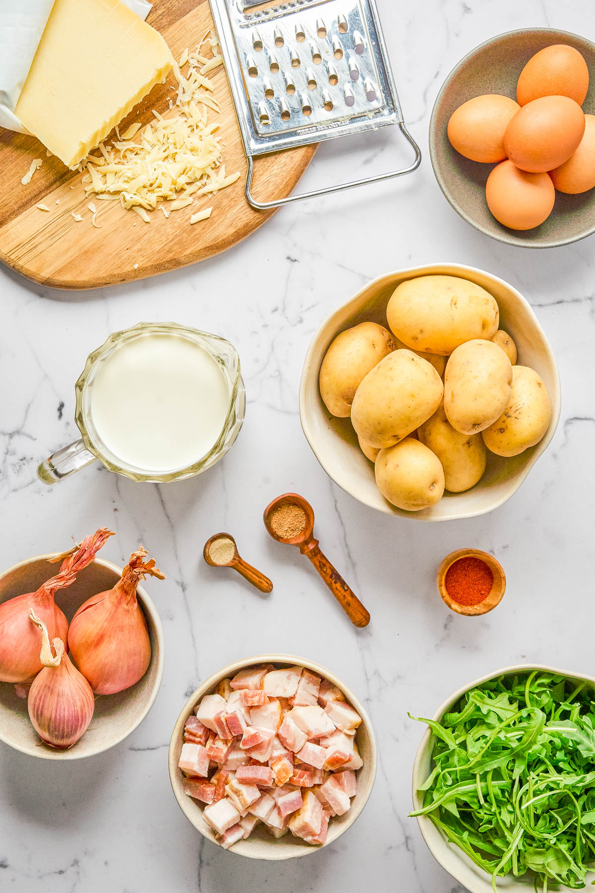 Various ingredients on a white surface, including potatoes, eggs, milk, cheese, shallots, diced bacon, arugula, spices, and a grater.