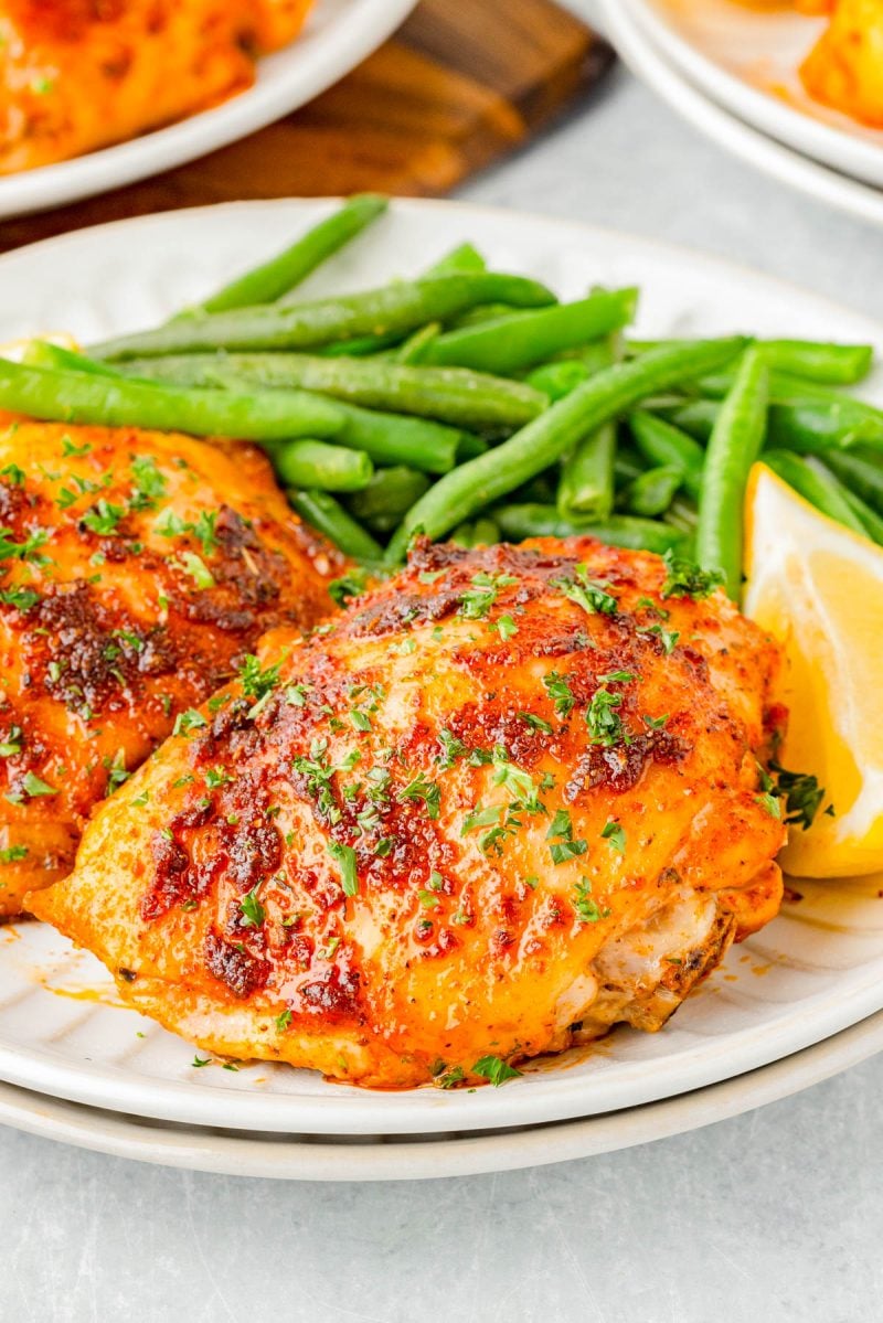 Two baked chicken thighs garnished with herbs, served with green beans and a lemon wedge on a white plate.
