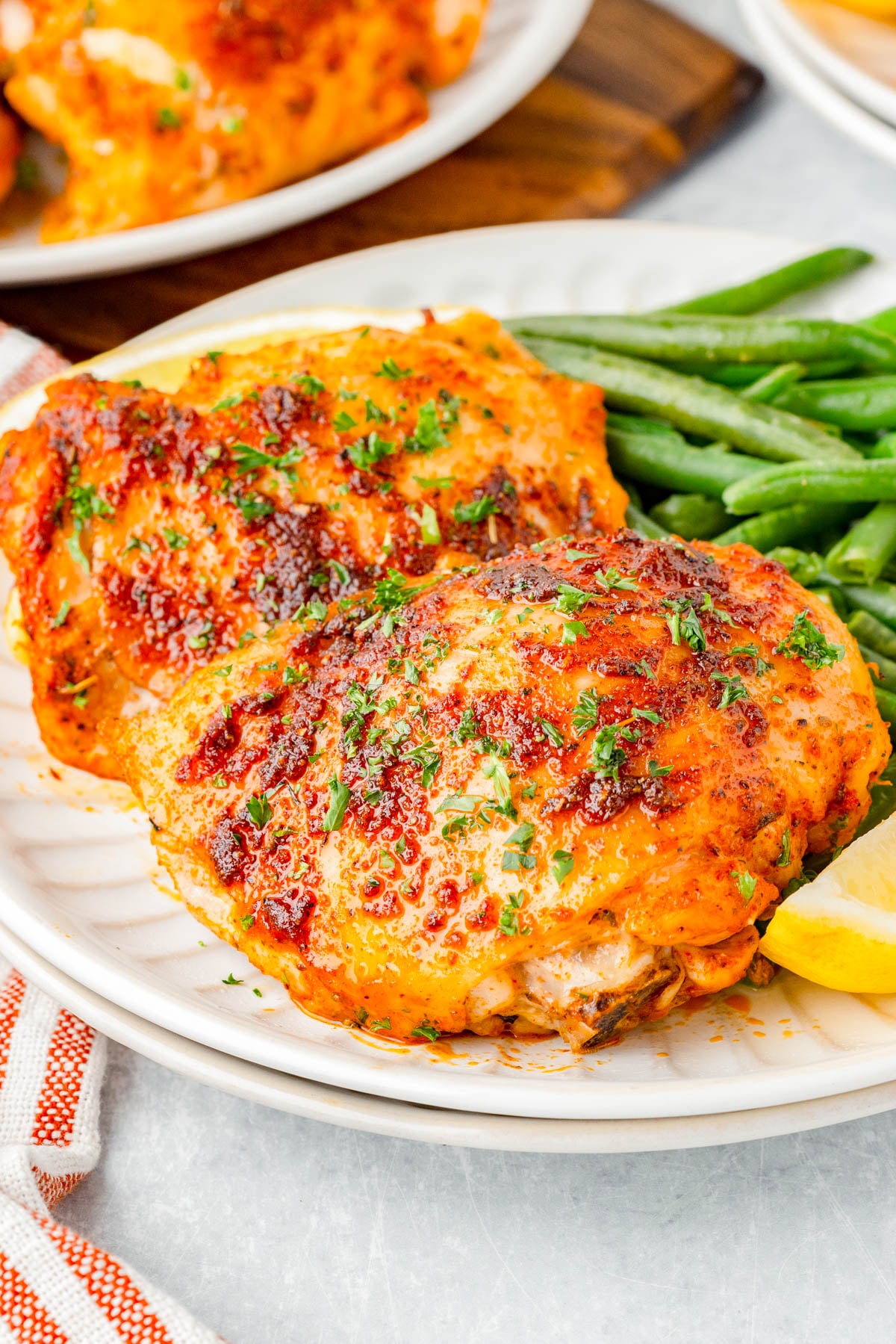 Two baked, seasoned chicken thighs served with green beans and a lemon wedge on a white plate.