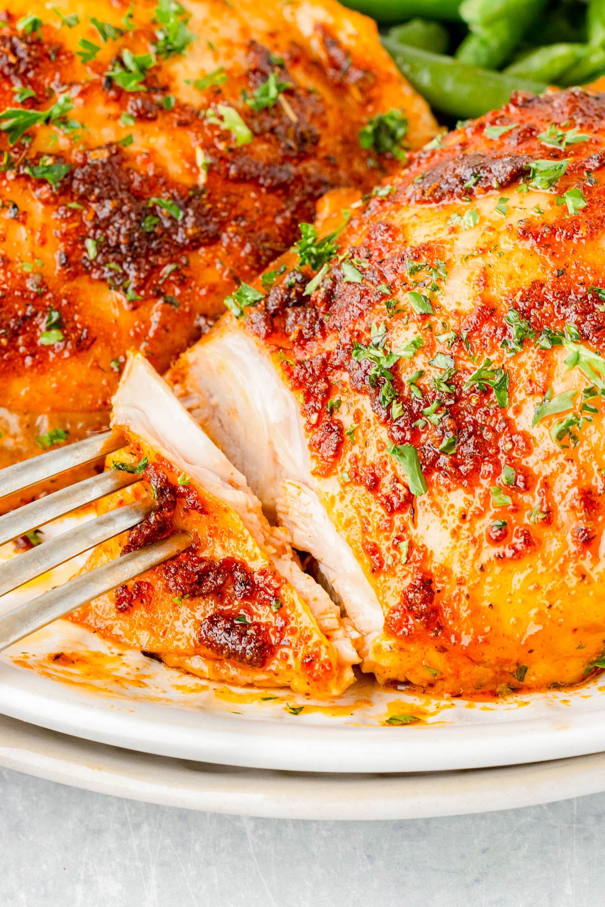 Close-up of a fork cutting into a seasoned, baked chicken breast garnished with chopped herbs on a white plate.