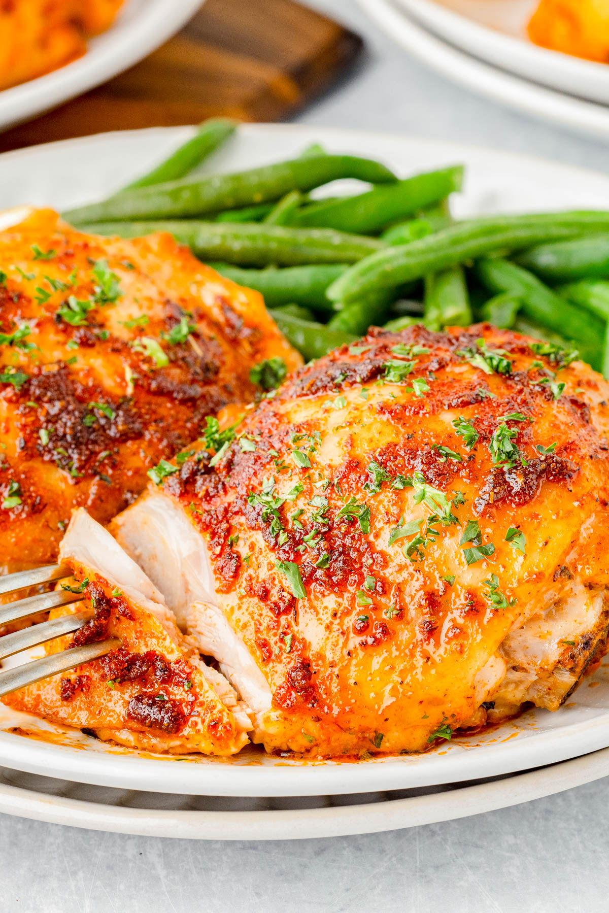 A plate of baked seasoned chicken breasts garnished with herbs, served with a side of green beans. A fork is cutting into one of the chicken pieces.
