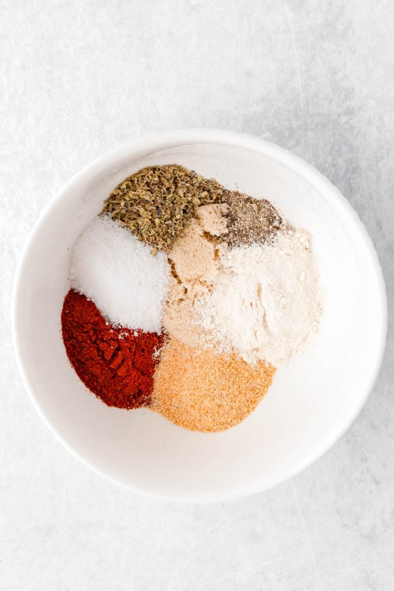 A white bowl containing six different spices and seasonings, arranged in separate piles on a light surface.