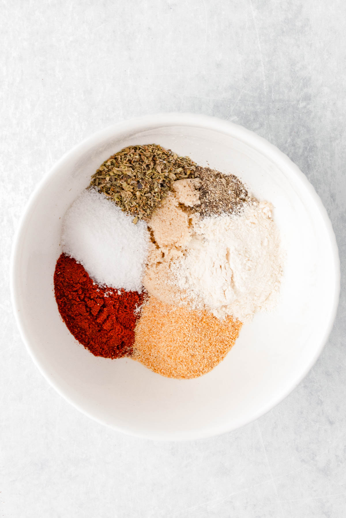 A white bowl containing six different spices and seasonings, arranged in separate piles on a light surface.