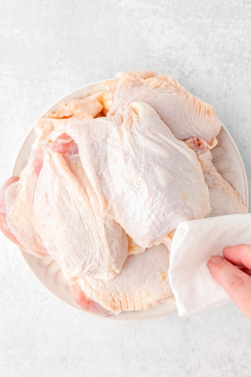 A hand pats raw chicken thighs with a paper towel on a white plate.