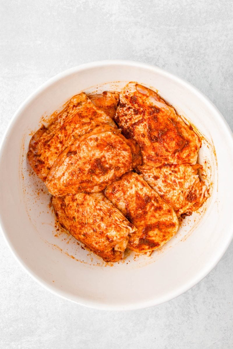 Raw chicken pieces coated in a red spice marinade sit in a white bowl on a light surface.