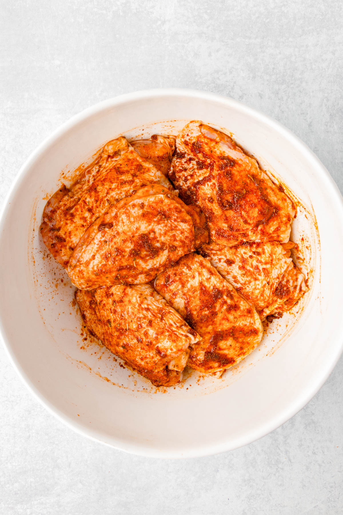 Raw chicken pieces coated in a red spice marinade sit in a white bowl on a light surface.