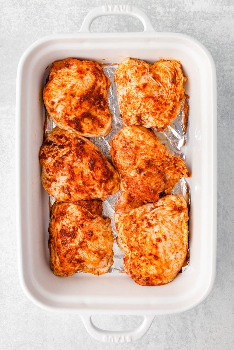 Six raw, seasoned chicken breasts arranged in a single layer on foil in a white rectangular baking dish.