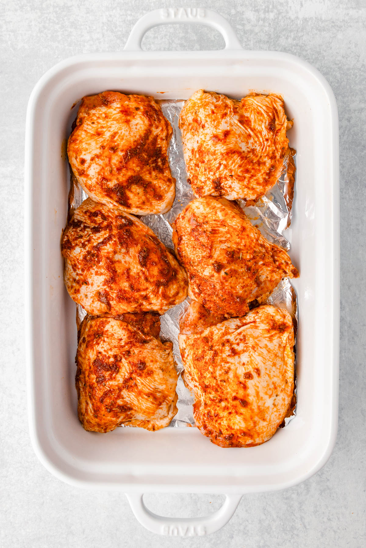 Six raw, seasoned chicken breasts arranged in a single layer on foil in a white rectangular baking dish.
