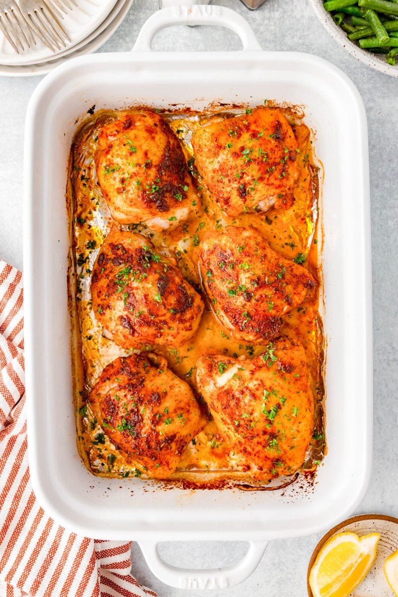 Six seasoned, baked chicken thighs sit in a white rectangular baking dish, garnished with chopped herbs and surrounded by juices.