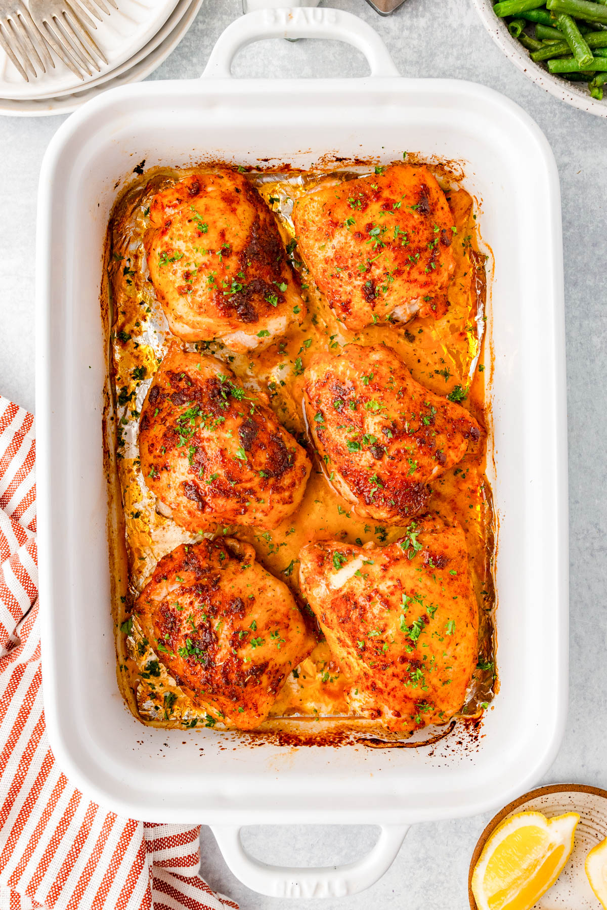 Six seasoned, baked chicken thighs sit in a white rectangular baking dish, garnished with chopped herbs and surrounded by juices.