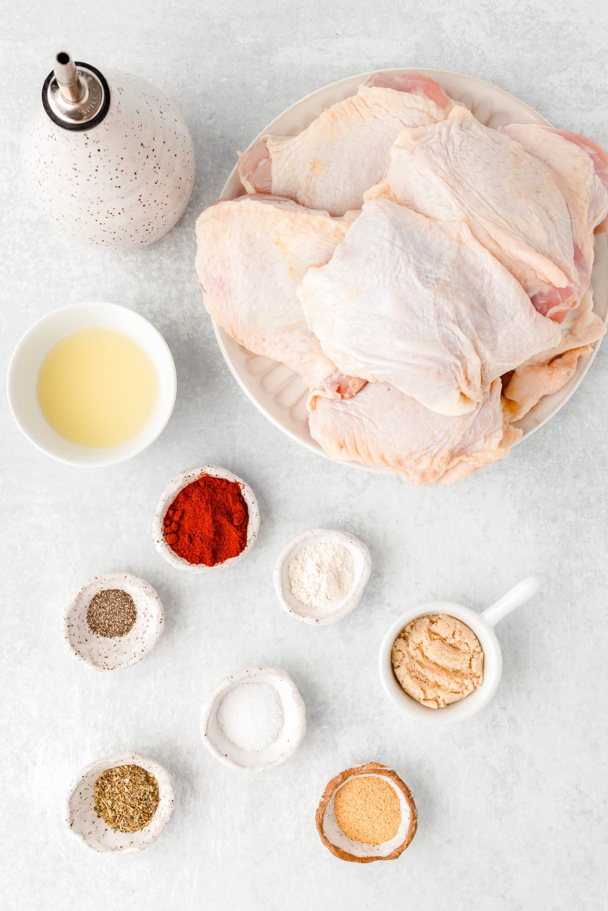 Raw chicken thighs on a plate with small bowls of oil, flour, brown sugar, paprika, black pepper, salt, Italian seasoning, and garlic powder arranged on a white surface.