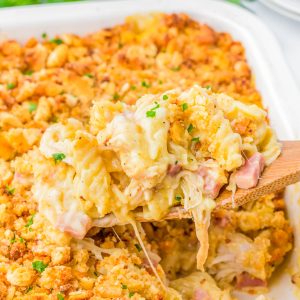 A wooden spoon lifts a serving of cheesy pasta bake with ham and a golden breadcrumb topping from a casserole dish.