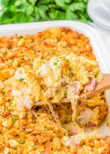 A wooden spoon lifts a serving of cheesy pasta bake with ham and a golden breadcrumb topping from a casserole dish.
