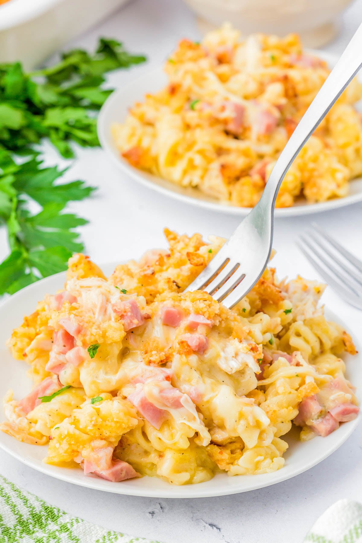 A fork is lifting a bite of cheesy pasta casserole with ham from a white plate, with another serving and parsley in the background.