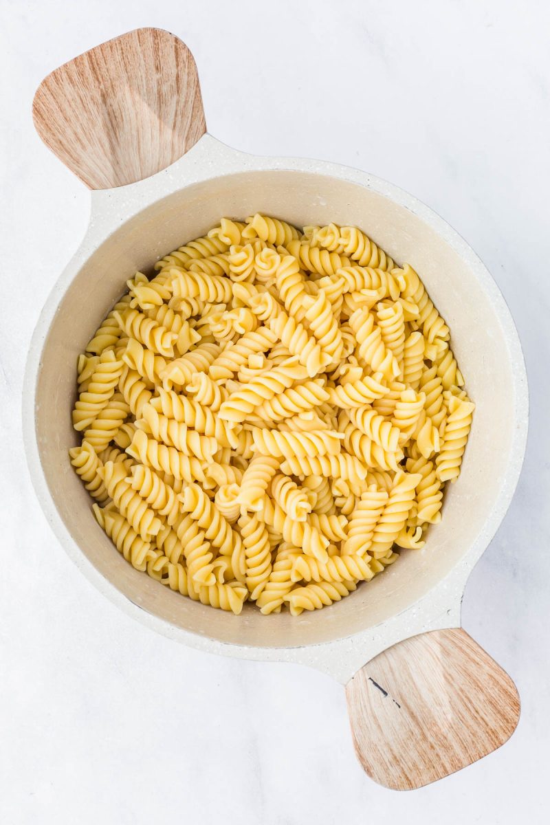 A white pot with wooden handles filled with cooked rotini pasta, viewed from above on a light surface.