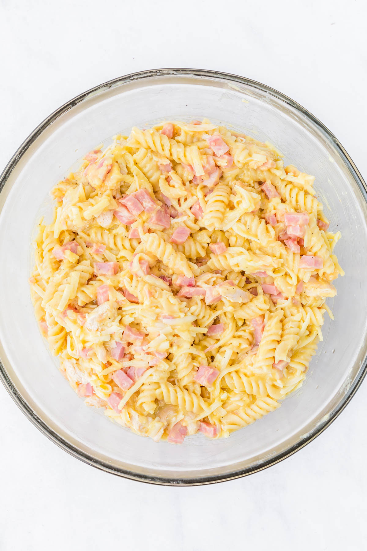 A glass bowl filled with rotini pasta mixed with diced ham and shredded cheese on a white surface.