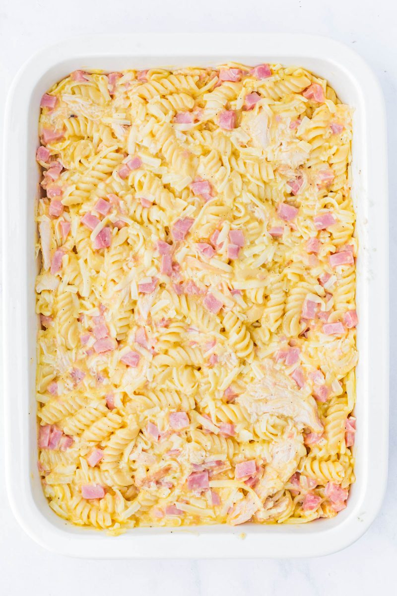 A white baking dish filled with cooked rotini pasta mixed with small pieces of ham and chicken in a creamy sauce.