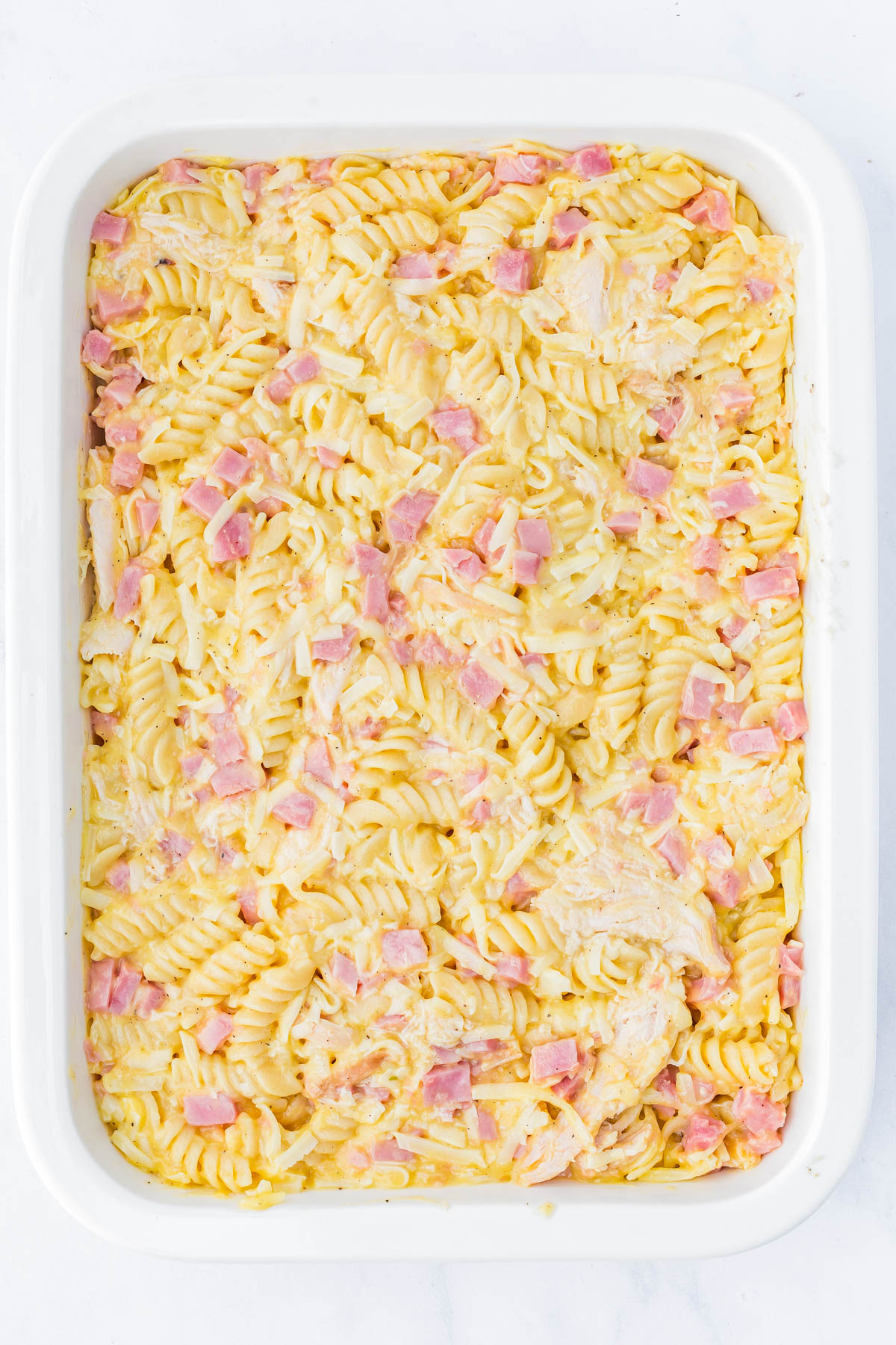 A white baking dish filled with cooked rotini pasta mixed with small pieces of ham and chicken in a creamy sauce.