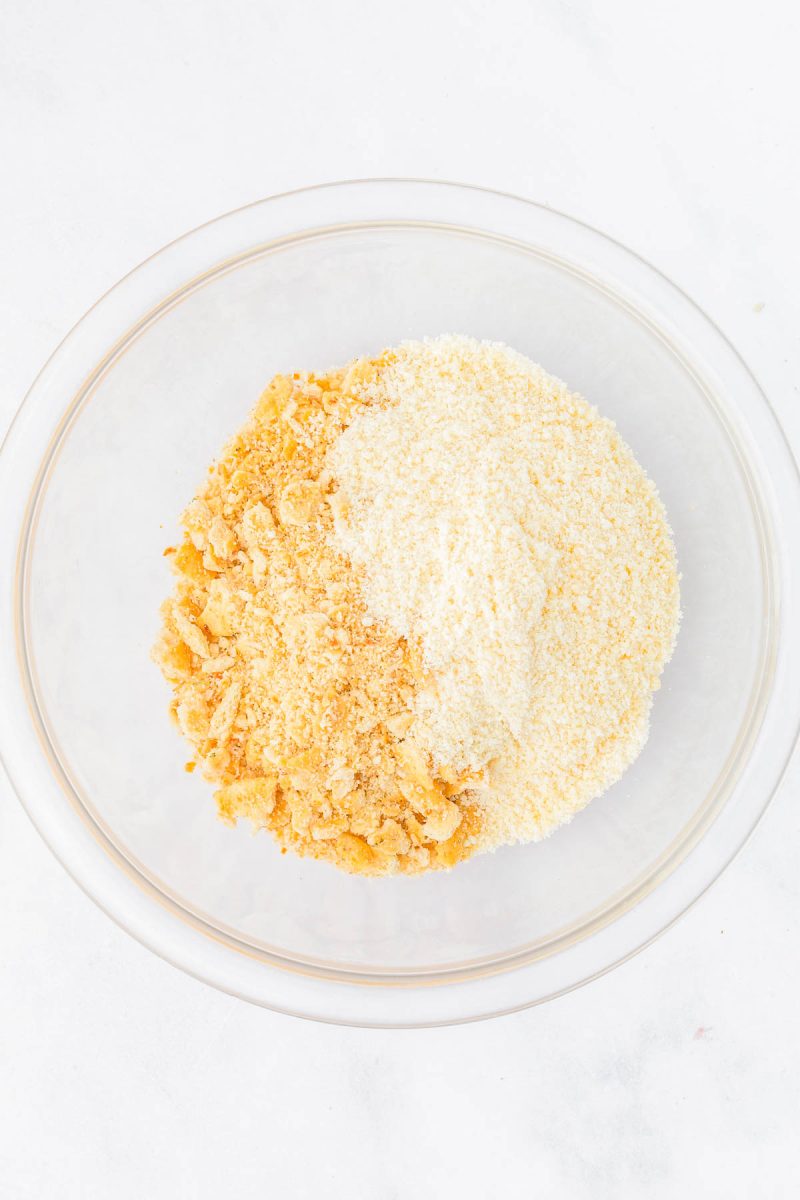 A glass bowl containing separate piles of crushed crackers and breadcrumbs on a white surface.