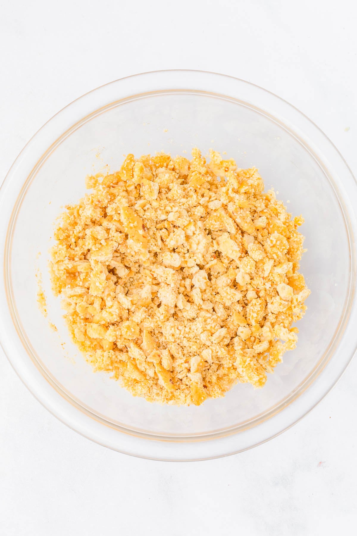 A clear glass bowl containing a crumbly mixture of crushed crackers or cookies on a white surface.
