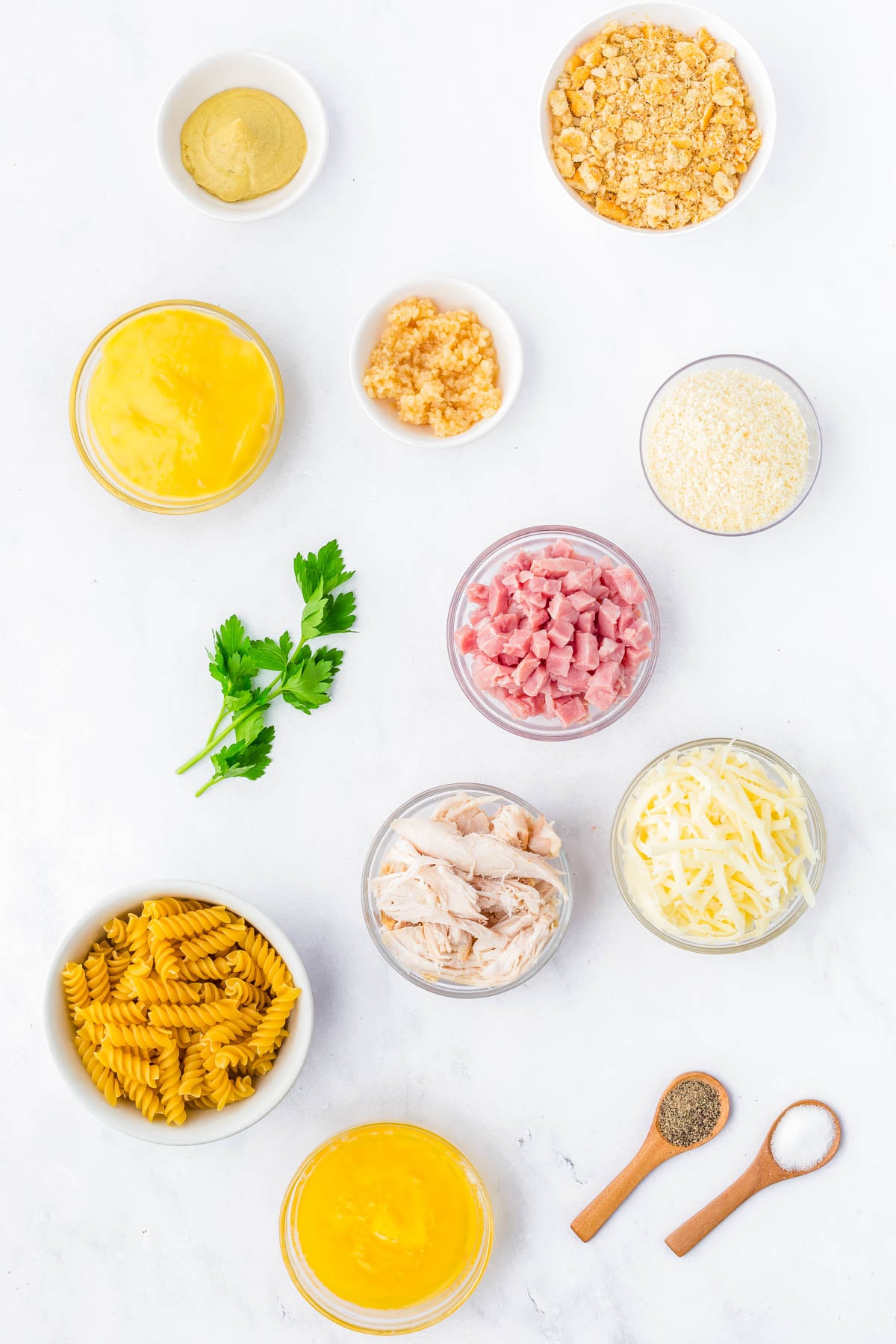 Top-down view of ingredients for a casserole, including pasta, shredded chicken, ham, shredded cheese, crushed crackers, breadcrumbs, mustard, garlic, eggs, parsley, salt, and pepper.