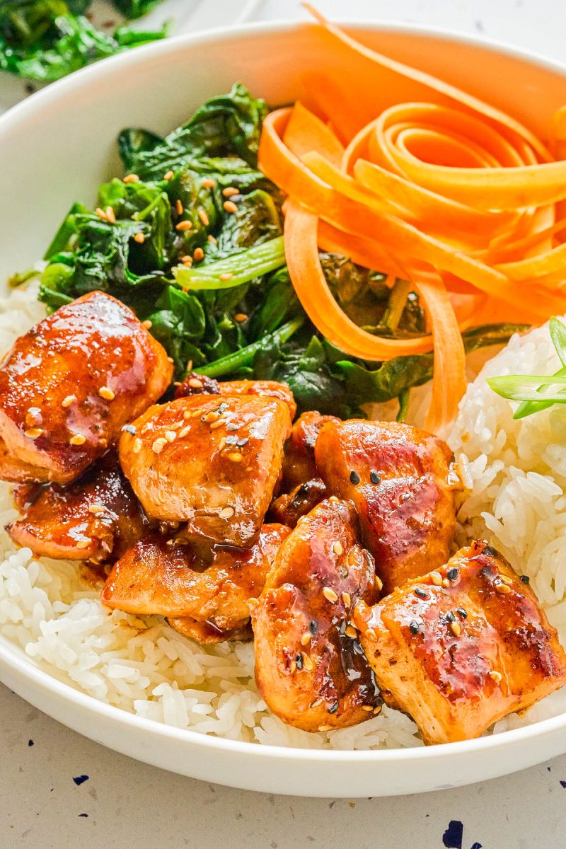A bowl of white rice topped with glazed salmon pieces, sautéed greens, and carrot ribbons.