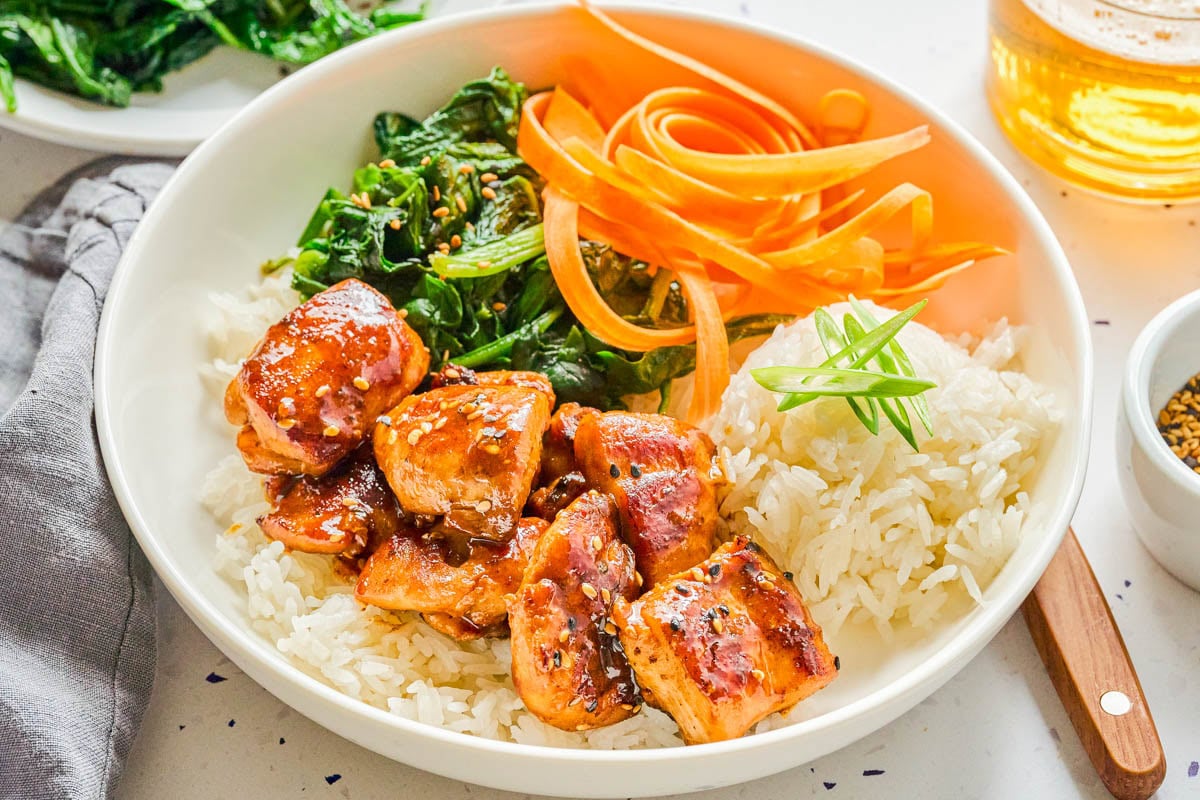Bowl with white rice, glazed chicken pieces, sautรฉed greens, carrot ribbons, and sliced green onions on top.