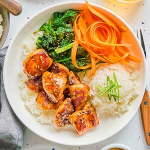 A white plate with rice, glazed chicken, sautéed greens, and carrot ribbons, garnished with sesame seeds and green onions. A drink and seasonings are beside the plate.