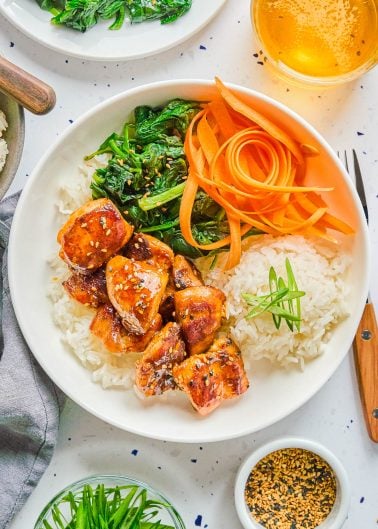 A white plate with rice, glazed chicken, sautéed greens, and carrot ribbons, garnished with sesame seeds and green onions. A drink and seasonings are beside the plate.