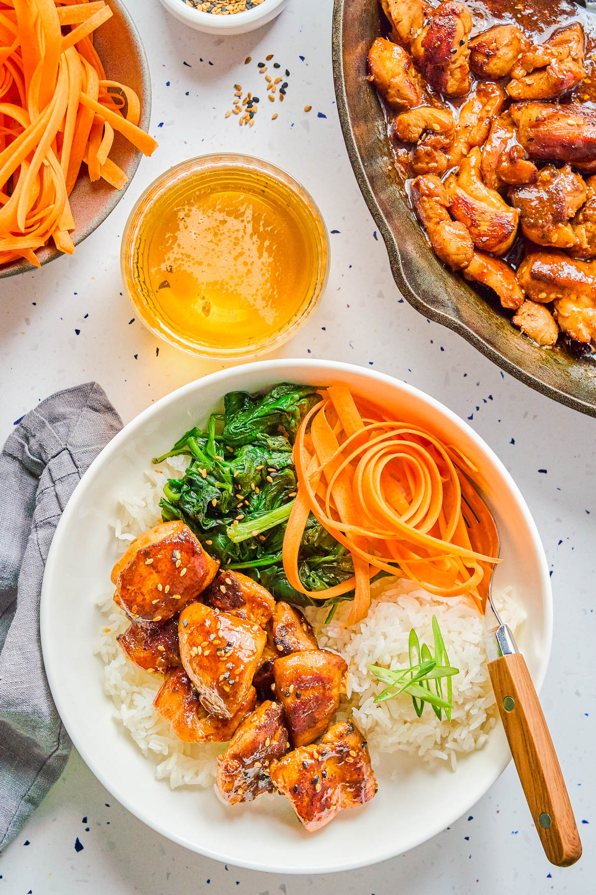A bowl of white rice topped with glazed chicken, sautรฉed greens, and raw carrot ribbons, with a fork. Surrounding items include raw carrots, a drink, and a pan of cooked chicken.