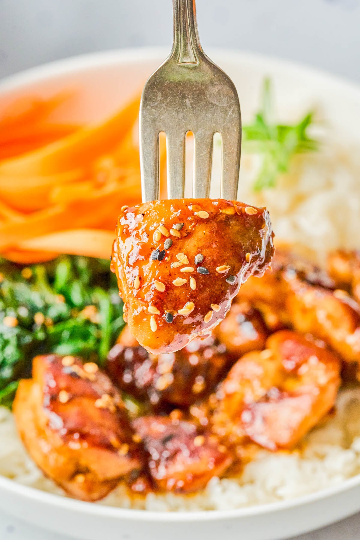 A fork holds a glazed piece of chicken sprinkled with sesame seeds above a plate with rice, spinach, and sliced carrots.
