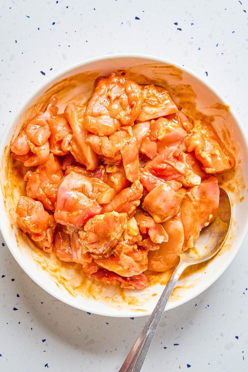 Raw, marinated chicken pieces in a white bowl with a metal spoon, placed on a white speckled surface.