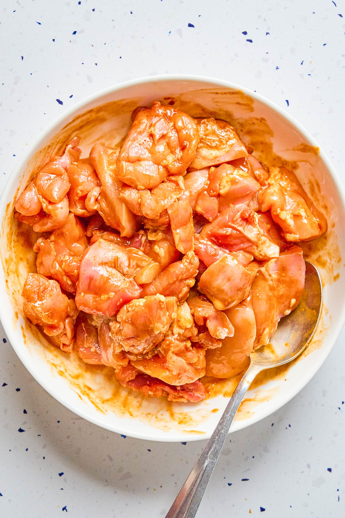 Raw, marinated chicken pieces in a white bowl with a metal spoon, placed on a white speckled surface.