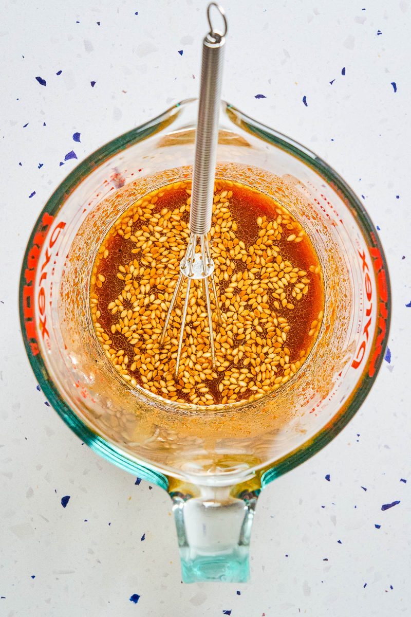 A glass measuring cup filled with a sesame seed and liquid mixture, with a metal whisk resting inside, on a white countertop.