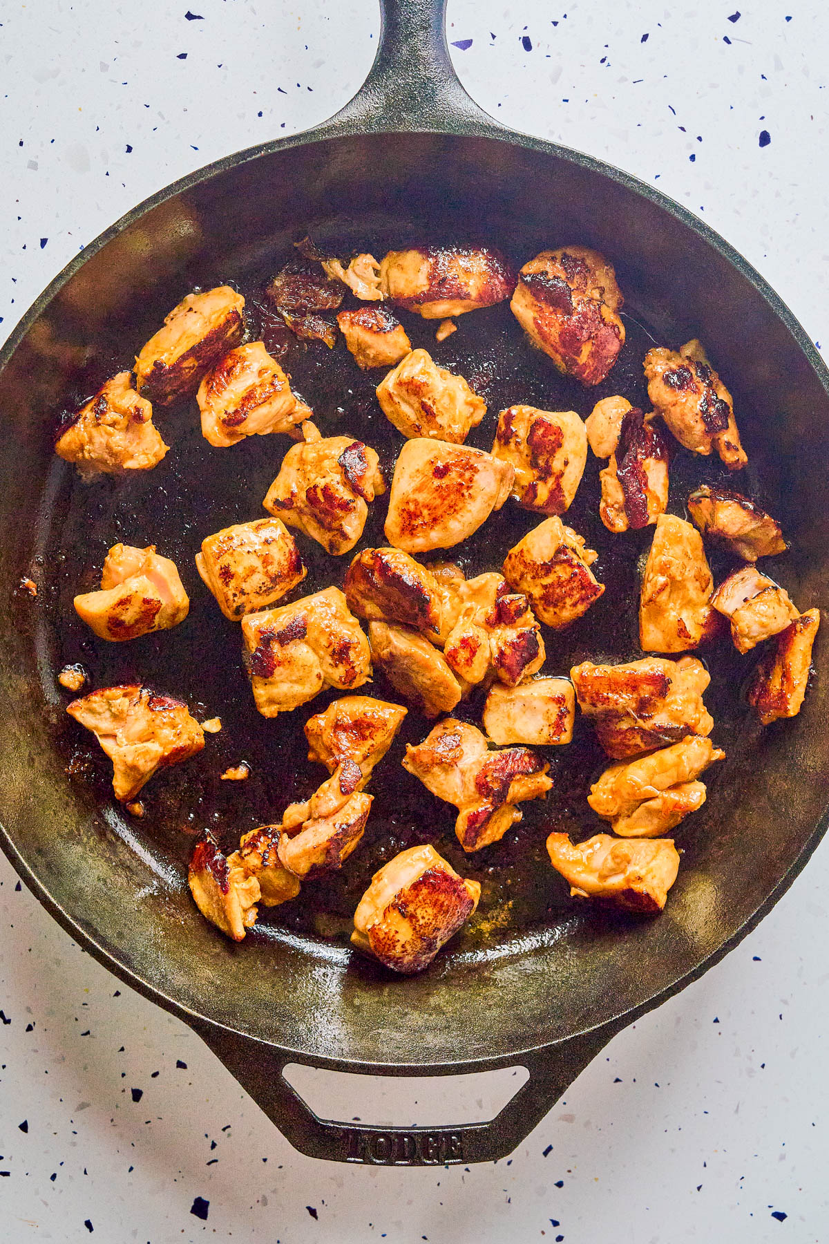 Chunks of browned chicken cooking in a cast iron skillet on a white, speckled countertop.