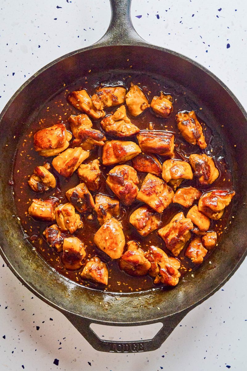 Chunks of cooked chicken coated in a brown sauce are in a black cast iron skillet on a white speckled surface.