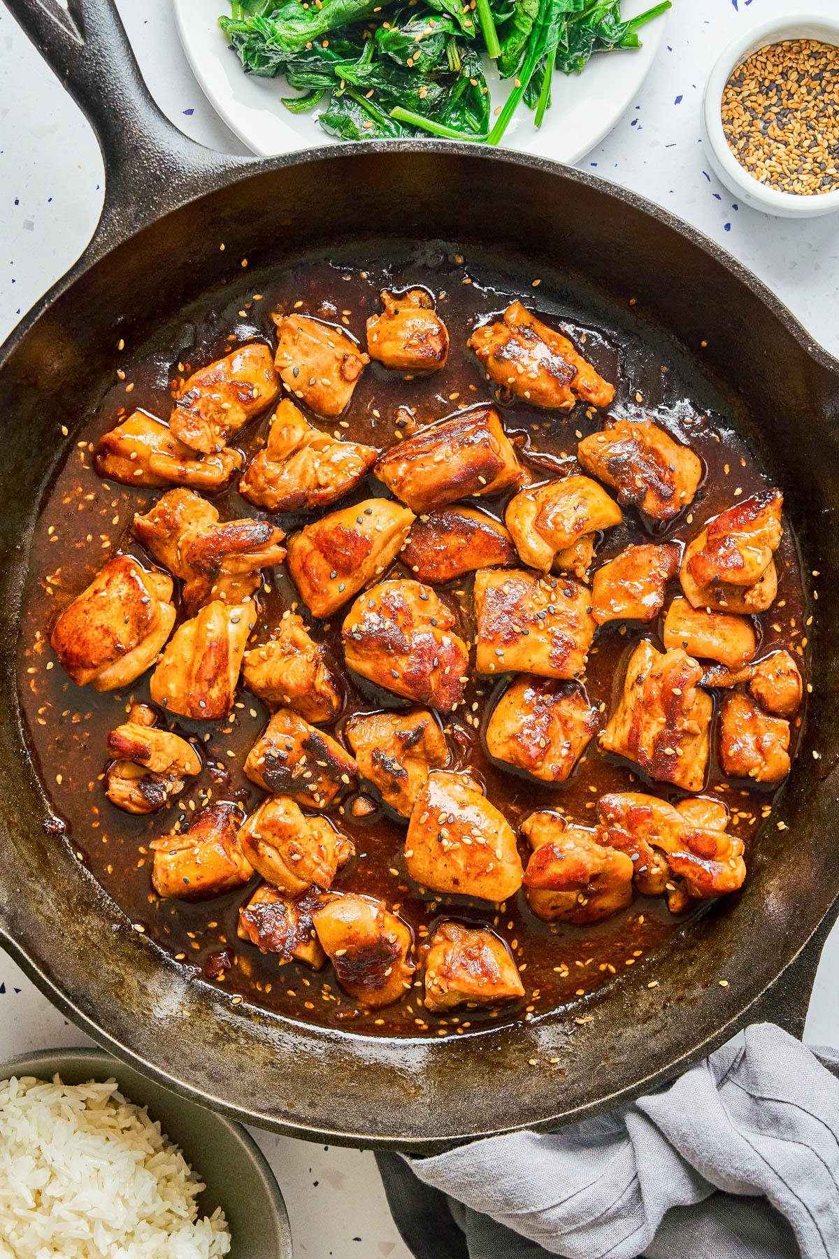Chunks of glazed chicken cooking in a cast iron skillet with sauce, surrounded by bowls of rice, sautรฉed greens, and sesame seeds.