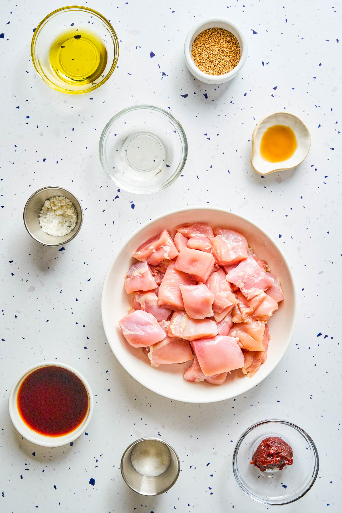 Overhead view of raw diced chicken in a bowl, surrounded by small bowls containing oil, sauces, seeds, salt, paste, and vinegar on a white speckled surface.