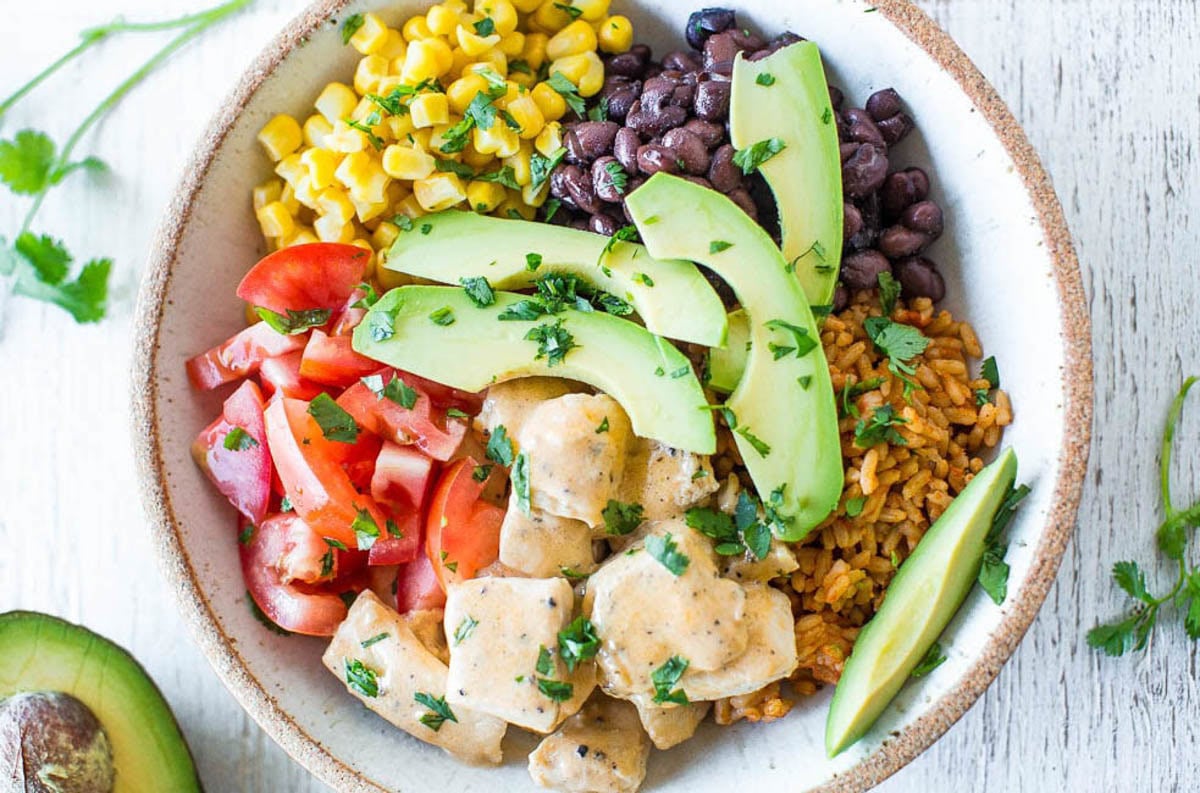A bowl containing diced tomatoes, corn, black beans, sliced avocado, seasoned rice, and creamy chicken pieces, garnished with chopped cilantro.