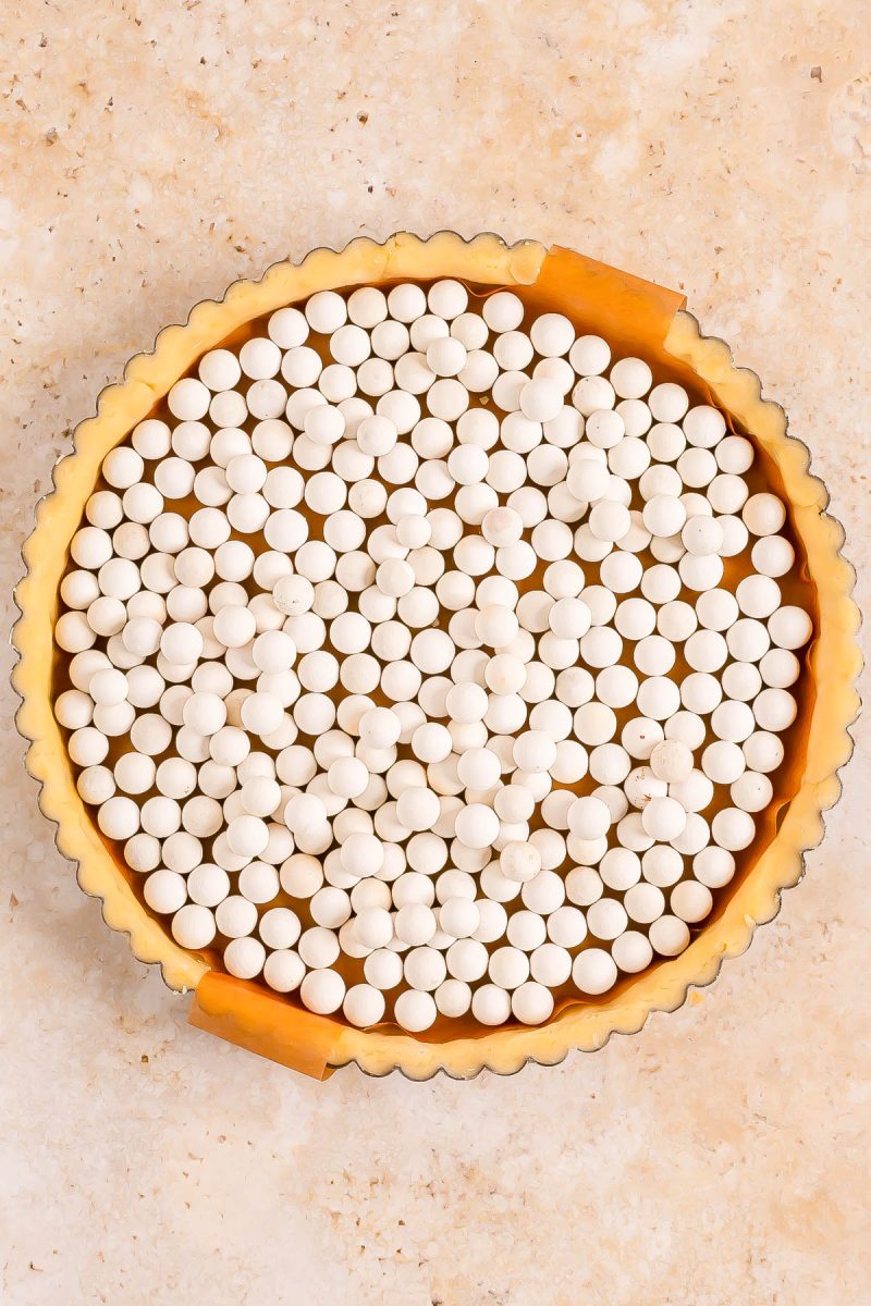 A tart pan filled with unbaked pie crust, lined with parchment paper, and topped with ceramic baking weights on a beige surface.