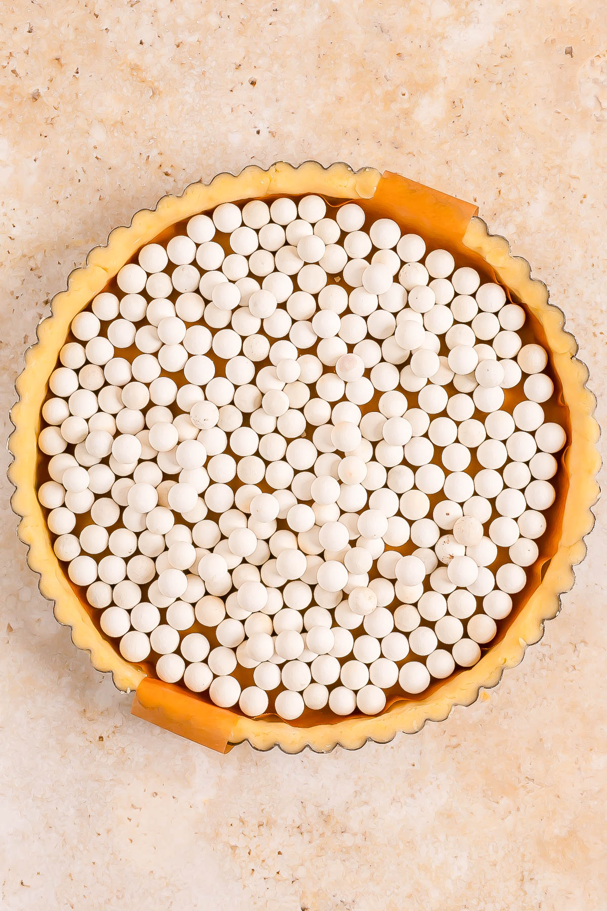 A tart pan filled with unbaked pie crust, lined with parchment paper, and topped with ceramic baking weights on a beige surface.