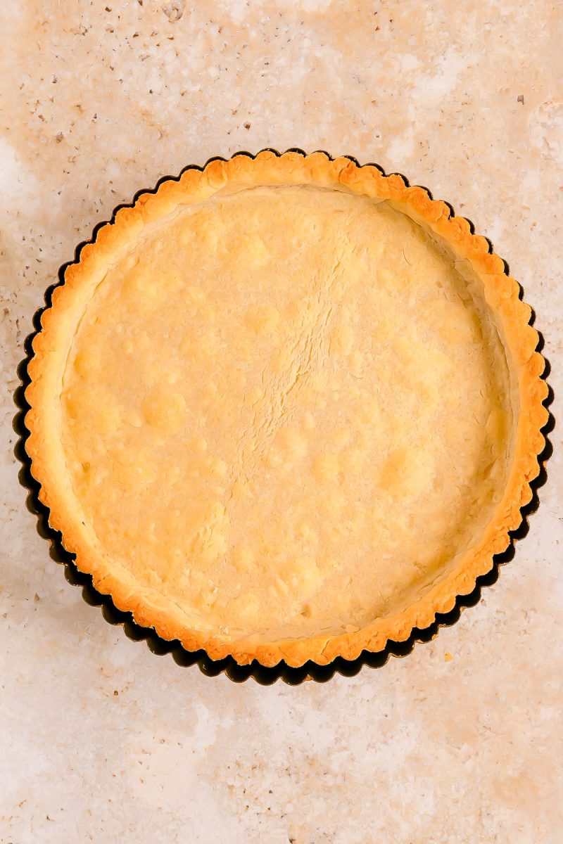 A baked, empty tart crust in a fluted tart pan rests on a light-colored, speckled surface.