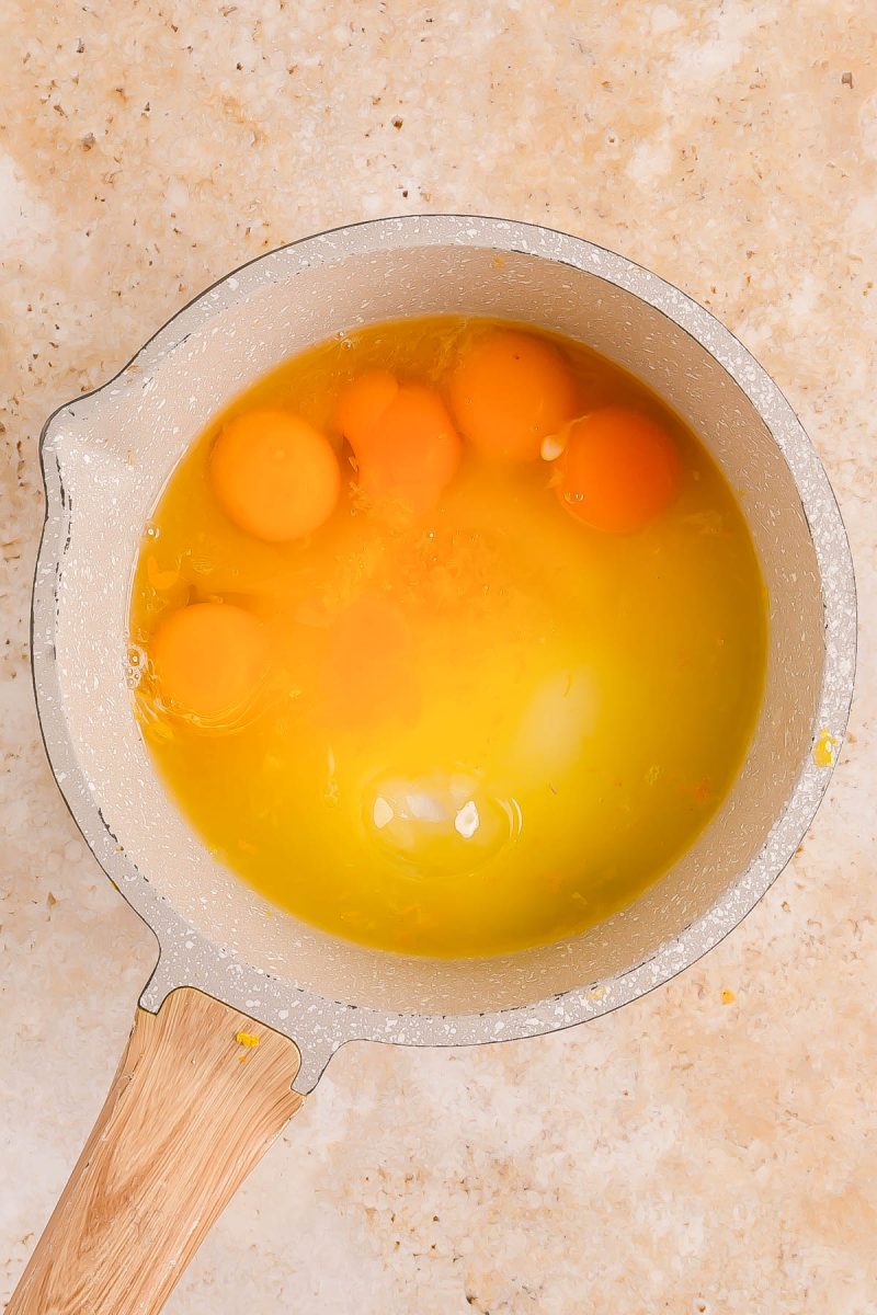 A saucepan containing several cracked raw eggs with intact yolks and whites, placed on a light-colored countertop.