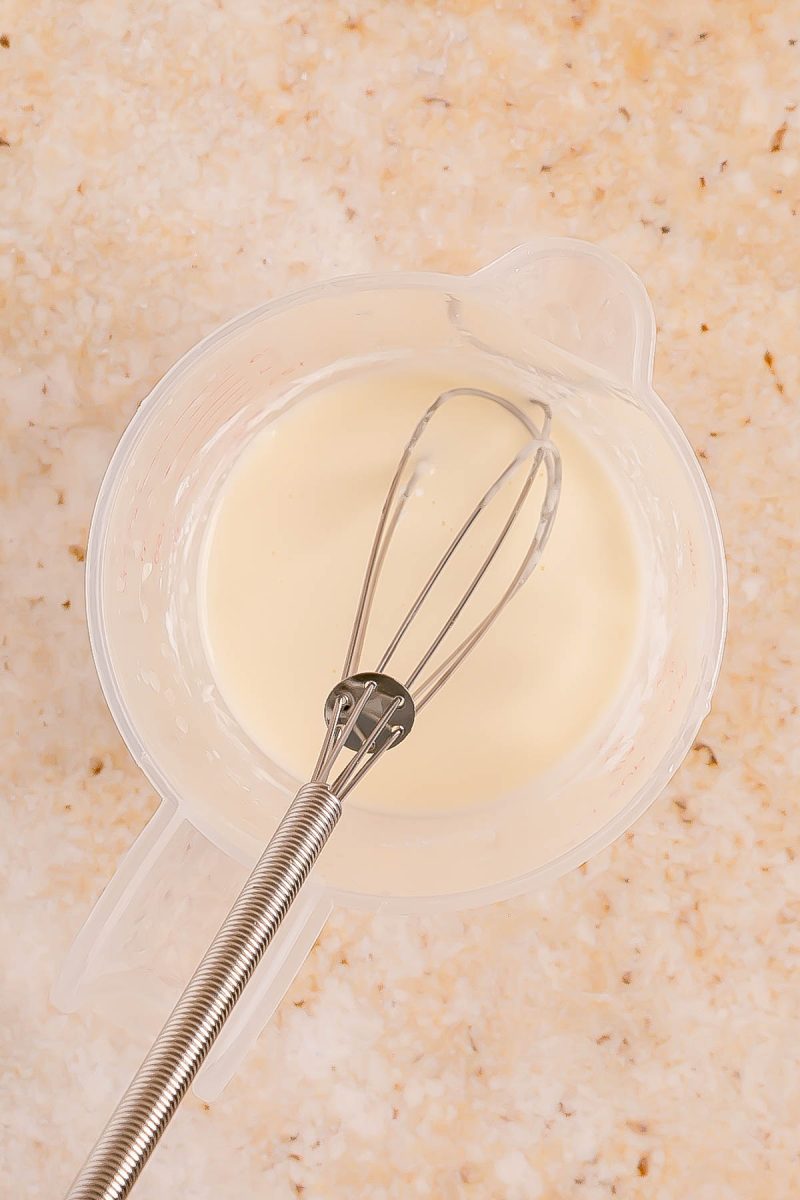 A whisk rests in a clear mixing bowl containing a pale, creamy liquid on a light marbled countertop.