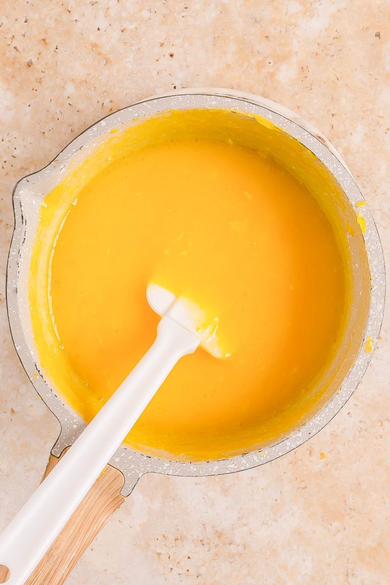 A saucepan filled with smooth yellow custard, with a white spatula resting inside, on a light-colored countertop.
