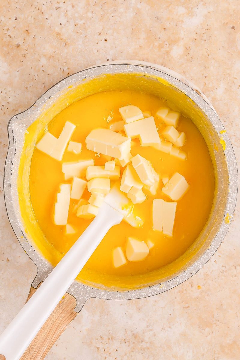 A saucepan filled with a yellow mixture and topped with chunks of butter being stirred with a white spatula on a beige countertop.
