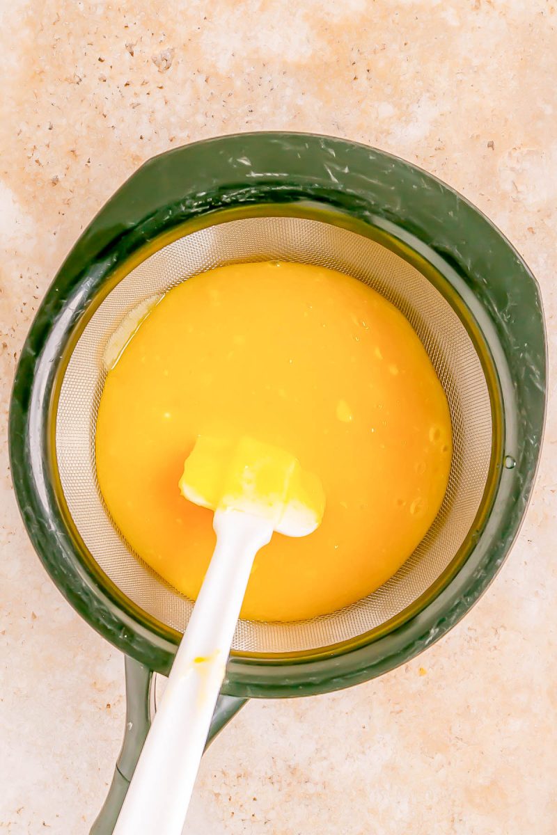 A mesh strainer containing yellow liquid and a white spatula, placed on a light countertop.