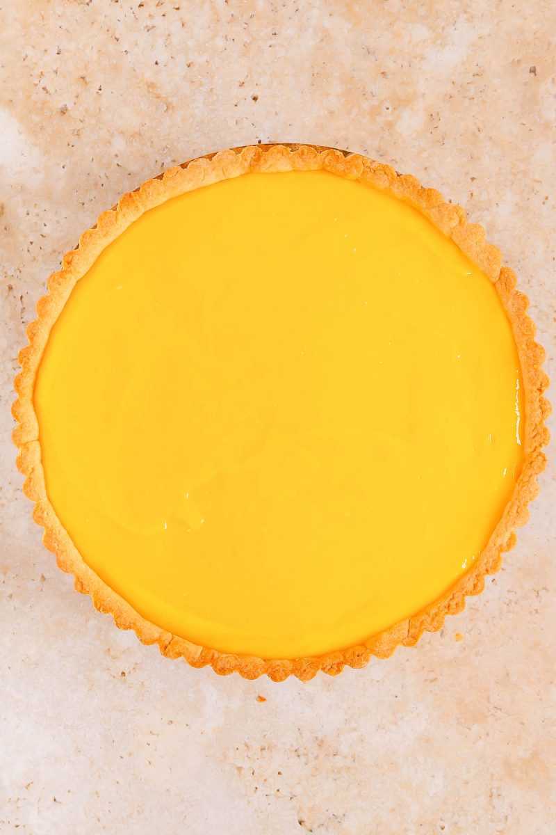 A round lemon tart with a smooth, glossy yellow filling and a golden brown crust on a light speckled surface.