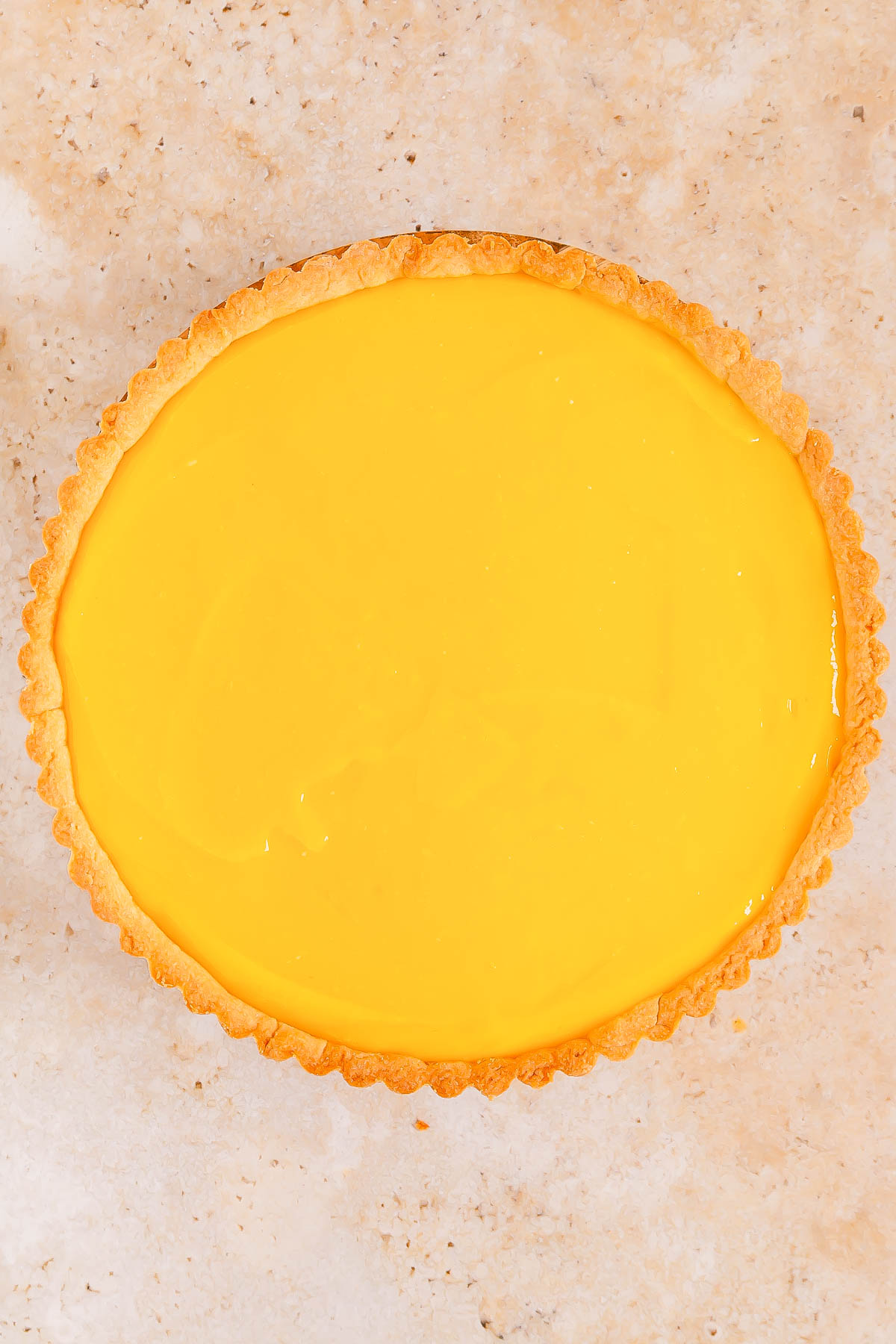 A round lemon tart with a smooth, glossy yellow filling and a golden brown crust on a light speckled surface.